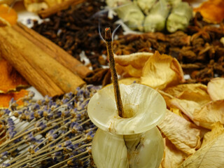Dried herbs, flowers, spice, orange peel and burning incense stick in an onyx incense holder, top view,  background. Ingredients for air freshening and relaxation.