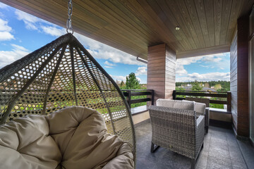 Naklejka premium A balcony features a comfortable hanging chair and a seating arrangement. Surrounding greenery and distant structures are visible under a bright sky with clouds.