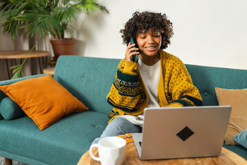 African American woman freelancer using laptop talking on phone at home office. Happy business girl having virtual meeting. Woman using phone laptop for communication working indoors. Remote work