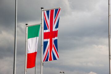Flags of Italy and the United Kingdom fluttering in the wind against a dramatic cloudy sky, symbolizing cultural connection and international relations in a globalized world