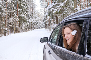 Young beautiful brunette teen girl looks out of the car window on the background of a snowy forest.  A smiling girl enjoys a winter car trip. Concept of weekend travel, leisure, a safe transportation.