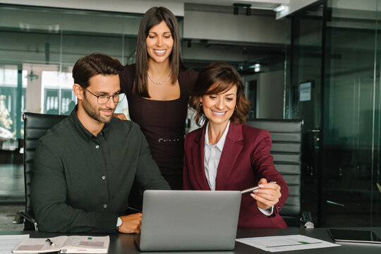 Smiling mature Latin hispanic business woman and European young team working using laptop computer in office. Diverse colleagues coworkers group of professional business people discussing project