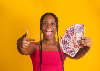 An excited young blacj african woman with braided hair holds a fan of 200 Ghanaian Cedi notes against a bright yellow background. She points toward the money with a joyful, open-mouthed expression of 