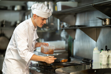Young man restaurant employee is busy preparing dish in kitchen. Worker roasts food in frying pan, cooks soup and performs auxiliary tasks..