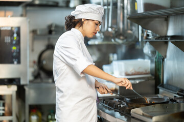 Girl restaurant employee is busy preparing dish in kitchen. Worker roasts food in frying pan, cooks soup and performs auxiliary tasks.