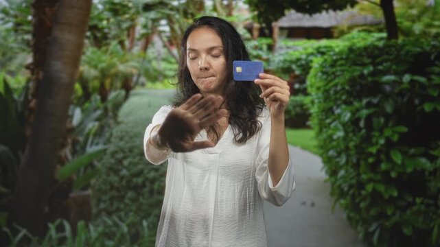 Young hispanic woman holding creditcard with hand raised in stop gesture on tropical garden path near hedges and palm trees; privacy refusal.