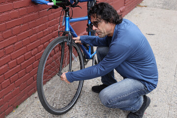 man in sunglasses checking on his bicycle (tire pressure bike maintenance) casual clothes commuter brick wall background urban cycling cyclist biker rider road mountain touring brakes handlebars nyc u