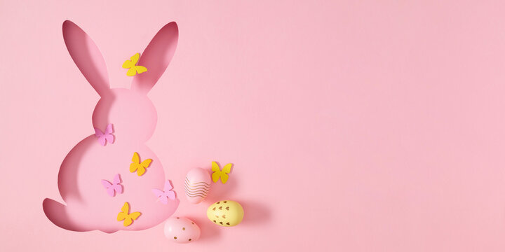 Stylish spring decor with a cut-out paper bunny silhouette, decorative eggs, and yellow butterflies on a soft pink background. Easter composition with bunny and eggs.
