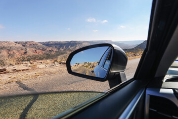 Road trip through desert landscape. View from car window with side mirror reflection. Scenic canyon road under blue sky. Travel freedom concept with open highway and rocky terrain in warm daylight.