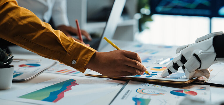 A human hand works with a robot's hand to analyze charts and graphs on a table, highlighting the fusion of technology and business strategies in modern workplaces.Noogenesis - Powered by Adobe
