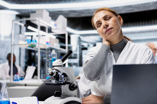 Exhausted scientist in lab suffering neck ache while documenting clinical trial outcomes on laptop. Fatigued researcher massaging stiff muscles in lab while updating database system