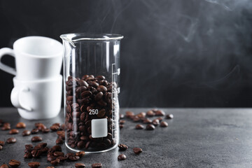 Coffee beans in glass beaker, cups and smoke on grey table, closeup. Space for text