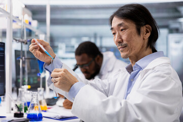 Close up of biochemist using pipette to transfer chemical sample into vial containing blue solution at laboratory bench. Asian research lab employee does pipetting procedure with assay liquid