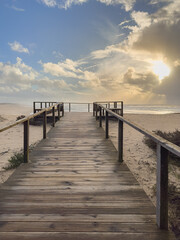 Obraz premium A rustic wooden boardwalk leads centrally across the sandy dunes of Furadouro beach Ovar Portugal to a platform overlooking the sea, illuminated by a brilliant sun breaking through dramatic clouds