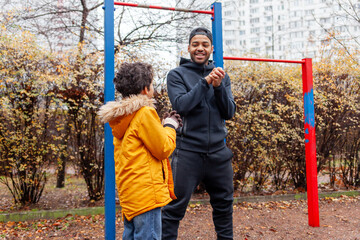 Obraz premium African American man and his son exercise and do exercises outdoors on playground, teenager stretches with his father on exercise equipment