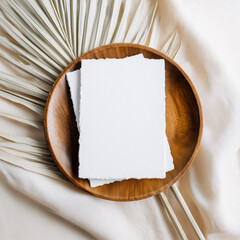 Minimalist stationery mockup with blank textured card on a wooden plate, styled with a dried palm leaf and soft fabric backdrop