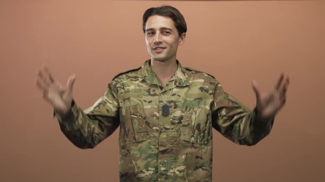 Man in camouflage uniform with hands raised and palms visible showing dogtag against peach studio backdrop; calm duty.