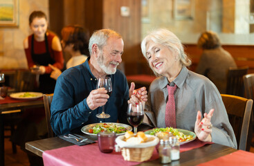 Senior spouses in cafe, chat and joke, flirting, eat and drink wine, spend time. Date, romantic meeting, nice time together. Atmosphere of cafe, restaurant conducive communication