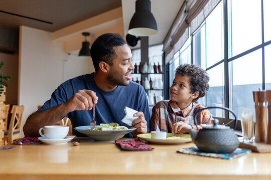 African American man and his son are having lunch in cafe at table, teenager and his father are sitting in restaurant and eating and drinking coffee