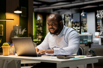 Black business consultant working late at night on proofreading documents, writing important...