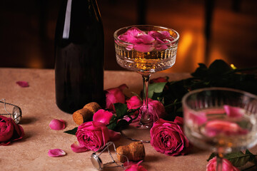 Obraz premium A delicate crystal champagne glass with golden liquid and rose petals, placed near a dark bottle, surrounded by pink roses and corks on a soft-lit table.