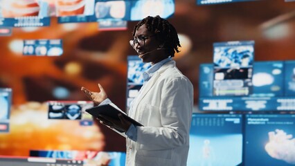 Cheerful woman showing futuristic medical visuals on digital videowall during conference. Jolly African american doctor presenting medical research data and 3D anatomy models dashboards, camera B