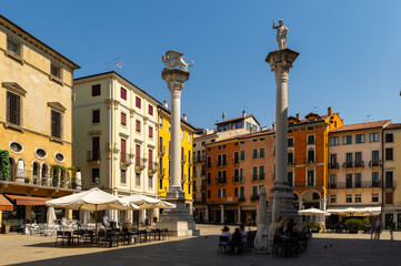Naklejka premium Two historic stone columns with sculpted figures rising in center of Piazza dei Signori in Vicenza, Italy, surrounded by colored facades of residential buildings and shaded seating areas on summer day