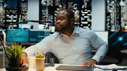 Black young businessman finishing tasks at midnight and closing his lamp, leaving office being overburdened and fatigued after doing overtime for complex project. Locking up.