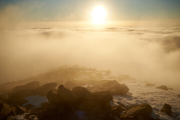 Sunrise over snow-covered mountain peak. Warm golden light of rising sun bathes clouds in soft hues of orange and pink. Snow and scattered rocks in foreground add texture and depth to scene.