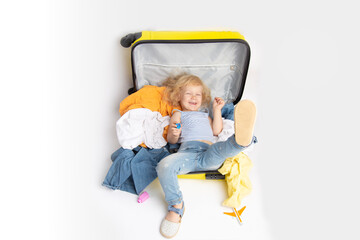 funny smiling curly baby girl lying in yellow suitcase with many clothes in white background. traveling with kids concept. banner with copy space. funny child