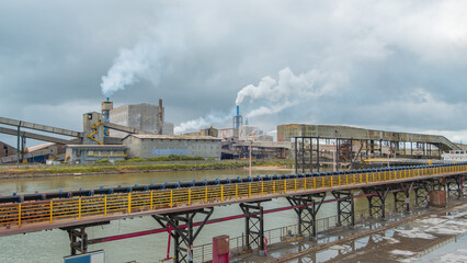 Industrial facility with smoking chimneys located on a riverbank, featuring long conveyor belts and steel structures under a cloudy sky at an automated plant
