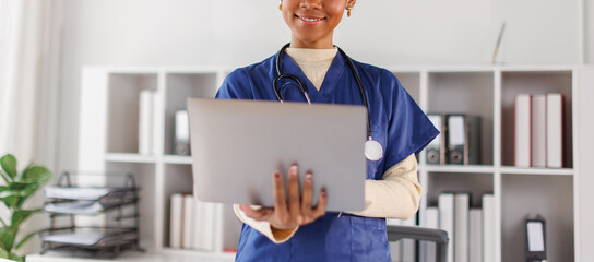 Medical professional in blue scrubs working on laptop, African american in afro hair smiling while...