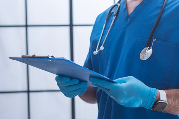 Male doctor wearing blue medical scrubs and stethoscope, holding a blue clipboard with patient...