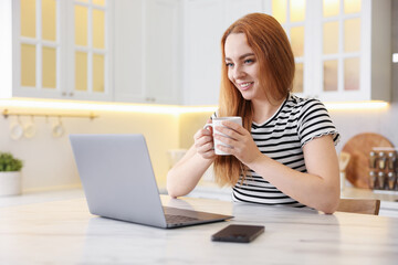 Woman having refreshing coffee while working on laptop at table in kitchen. Space for text
