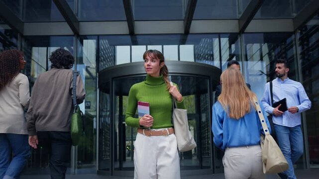 Tracking shot of a caucasian millennial woman in a green sweater walking out of a modern office entrance as coworkers pass by, conveying confidence, career momentum and urban corporate life.
