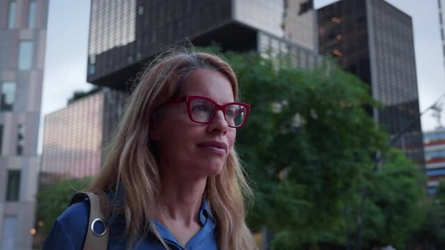 Confident businesswoman with red glasses walks through a modern office district at dusk, focused and professional in a blue shirt and backpack, conveying commute, career and urban lifestyle.