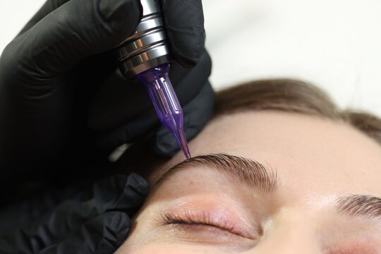 Beautician making permanent eyebrow makeup to young woman against light background, closeup