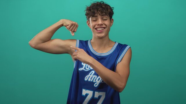 Teenage boy flexes bicep and points finger while smiling in studio wearing blue los angeles basketball jersey number 77; youthful pride.