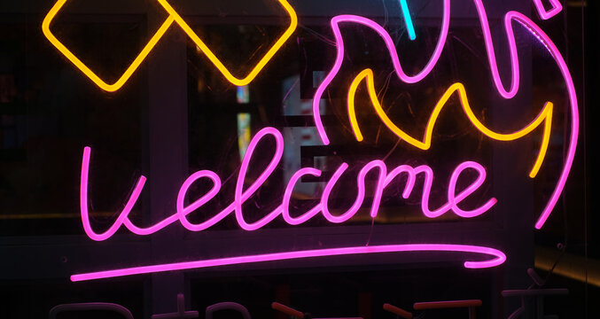 A glowing neon light sign mounted on a dark window, featuring the word Welcome in bright pink cursive typography