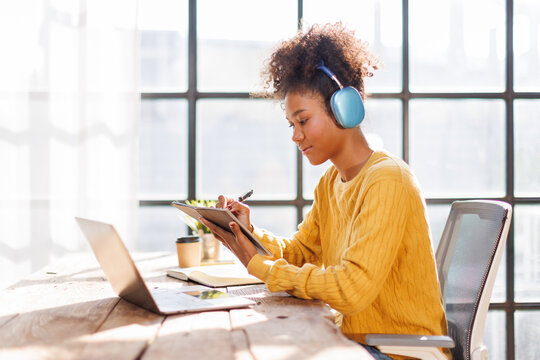 African american in afro hairwearing headphones using laptop in cafe, writing notes, attractive female student learning language, watching online webinar, listening audio course, e-learning education 