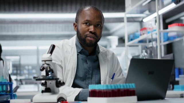 Hematology specialist in lab looking at blood sample in test tube, using laptop software. Epidemiologist using notebook program to analyze sanguine fluid specimen in vial, camera B