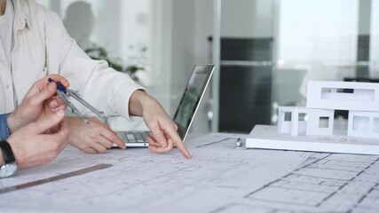Architects collaborating over blueprints and a white scale model, using ruler and caliper on a shared desk as they plan residential and urban building projects