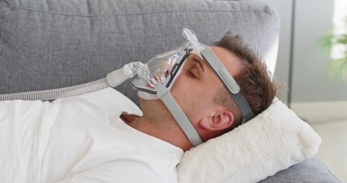 Man wears mask for sleep apnea therapy. A cpap oxygen interface supports breathing for a patient at home on a couch, illustrating healthcare and treatment comfort. Clear concept of safe sleep.