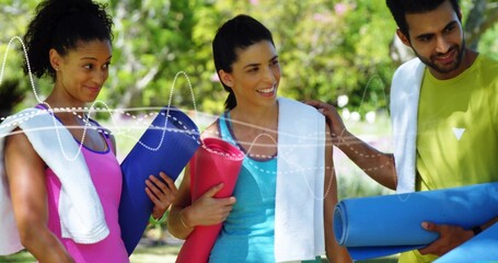 Standing three adults in athletic wear holding rolled yoga mats and towels in sunlit park, smiling
