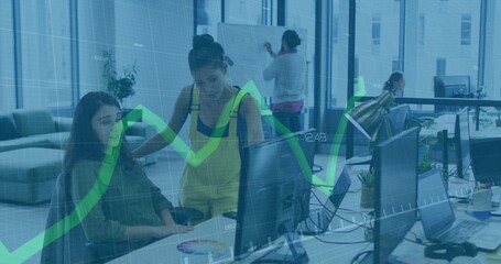 Working two women in yellow-top, leaning and pointing at monitor, open office, showing green graph