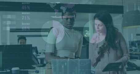 Viewing two women in pink blouse analyzing laptop with tablet and data overlays in open-plan office