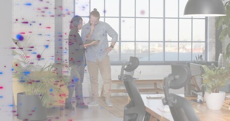 Reviewing tablet, two coworkers in dark blazer and rolled sleeves standing by harbor window, plants