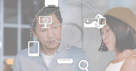 Working Asian colleagues in blue button-up and beige beanie examining AI icons in softly lit office