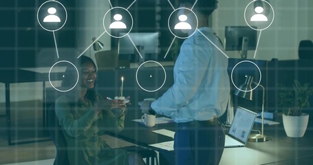 Toasting pair in workwear holding cupcake with lit candle at office desk laptop charts user icons