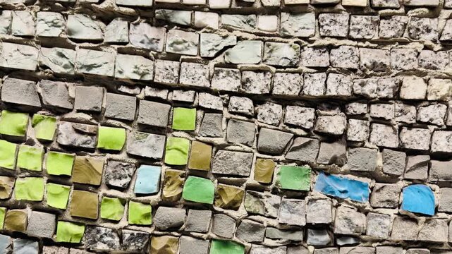 Vibrant stone mosaic wall closeup showing green and blue square tiles set in rough mortar, weathered grout lines, uneven beige stones, sunlit texture, urban exterior, decorative.